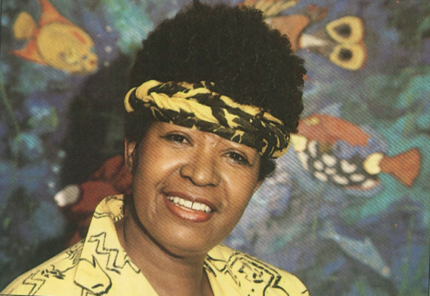 Foto de mulher negra sorrindo e olhando diretamente para a câmera. Ela tem cabelos curtos, escuros e crespos, olhos castanhos, veste camisa amarela com detalhes pretos e usa um lenço amarelo e preto enrolado e amarrado na testa. Atrás dela há uma ilustração de peixes coloridos em fundo azulado.