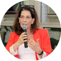 Foto de uma mulher branca ao microfone, falando e gesticulando com uma das mãos. Ela tem cabelos castanhos e lisos, na altura dos ombros, veste blusa branca e casaco vermelho e tem óculos sobre a cabeça. Suas unhas estão pintadas e ela usa anéis e relógio. Ao fundo, janelas de vidro.