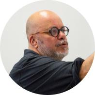 Foto de um homem de pele clara, careca e com barba grisalha falando. Ele está em semi-perfil com um braço levantado, usa óculos com armação redonda preta e camisa preta. O fundo é branco. 
