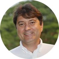 Foto de um homem de pele clara, olhos escuros e cabelos lisos, curtos e castanhos. Ele sorri e usa camisa branca. Ao fundo, há uma vegetação em desfoque. 