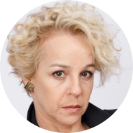 Foto de mulher branca de olhos castanhos e cabelos loiros, curtos e cacheados. Ela usa camisa preta e  brincos em formato quadrangular. O fundo é branco. 
