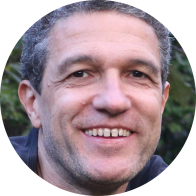 Foto de homem branco sorrindo. Ele tem olhos castanhos, cabelos curtos e grisalhos, usa camiseta azul escura e olha para a câmera. Ao fundo, folhagens.