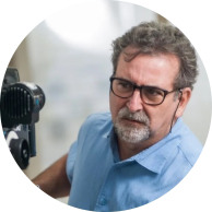 Foto de um homem branco de semi perfil manipulando uma filmadora. Ele tem cabelos curtos, lisos e grisalhos, olhos castanhos, cavanhaque grisalho. Usa óculos de armação escura e retangular e veste camisa azul. O fundo da imagem é claro e desfocado.