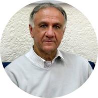 Foto de um homem branco com  cabelos curtos, lisos e grisalhos. Ele usa uma blusa cinza clara por cima de uma camisa branca. O fundo é uma parede branca com textura.