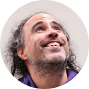 Foto de homem branco sorrindo e olhando para cima. Ele tem olhos castanhos, cabelos grisalhos e encaracolados, de tamanho médio, tem barba e bigode grisalhos e veste camisa com gola roxa. O fundo é bege.