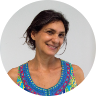 Foto de mulher branca sorrindo. Ela tem olhos castanhos, cabelos na altura das orelhas, lisos e castanhos. Ela está com a cabeça um pouco inclinada e usa uma blusa regata colorida. O fundo é branco.