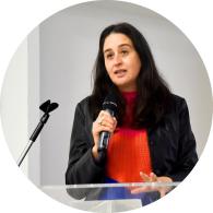 Foto de uma mulher branca segurando um microfone e falando. Ela tem cabelos longos, escuros e lisos, olhos castanhos, veste blusa laranja e blazer preto. A seu lado, um pedestal para microfone e à frente um púlpito transparente. O fundo da imagem é cinza.