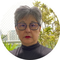 Foto de mulher amarela, com cabelos grisalhos, lisos e curtos. Ela olha diretamente para a câmera,  veste uma blusa preta , usa óculos de armação azul e batom vermelho. Ao fundo, um prédio, folhas de uma árvore e o céu claro. 