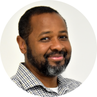 Foto de um homem negro que sorri em semi-perfil. Ele tem cabelos pretos e curtos, bigode e barba grisalhos e olhos castanhos escuros. Ele veste camisa xadrez sobre uma camiseta branca. O fundo é claro.