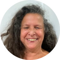Foto de mulher branca sorrindo. Ela tem cabelos compridos, ondulados e castanhos, com alguns fios grisalhos. Os olhos castanhos estão quase fechados. O fundo da imagem é claro. 