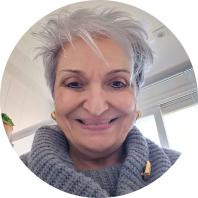 Foto de mulher branca com cabelos curtos, lisos e grisalhos. Ela está sorrindo diretamente para a câmera, veste um casaco de lã cinza e usa brincos dourados. No fundo, uma janela de vidro e parte do teto.