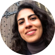 Foto de mulher branca sorrindo. Ela tem cabelos pretos, compridos e ondulados. Os olhos são castanhos e os lábios estão rosados. Ela usa uma blusa de gola alta marrom e, atrás dela, há uma parede de madeira. 