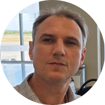 Foto de um homem branco, com olhos castanhos e cabelos levemente grisalhos. Ele veste uma camisa xadrez. Ao fundo, uma parede bege e uma porta de vidro.