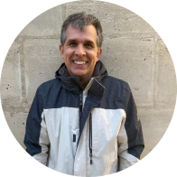 Foto de um homem branco sorrindo. Ele tem cabelos curtos, lisos e grisalhos e veste uma blusa azul marinho e branca. Atrás dele há uma parede de blocos de concreto.