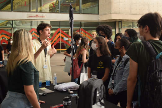 Foto de jovens conversando. Um homem branco, de cabelos curtos escuros e que veste camisa amarela, está gesticulando e falando para outros jovens. Do lado direito da imagem, cerca de cinco estudantes olham para ele. Na frente dessas pessoas, há uma mesa com uma toalha preta, copos de água e alguns brindes. A maioria das pessoas usa máscara.