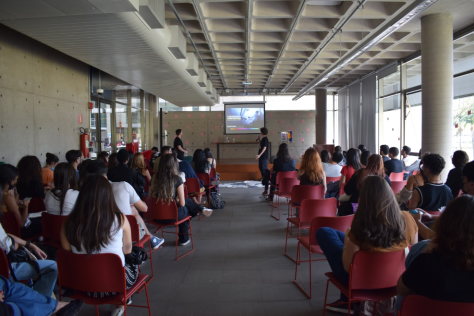Foto de uma palestra tirada do fundo da sala. Há diversos jovens de costas sentados em cadeiras vermelhas. As cadeiras estão dispostas para formar um corredor. Ao final desse corredor, no centro da imagem, está um homem segurando um microfone em frente a um telão com uma projeção. Ele é branco, tem cabelos curtos escuros e veste uma roupa preta. As paredes e o chão da sala são cinzas.