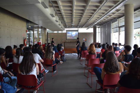 Foto de uma palestra tirada do fundo da sala. Há diversos jovens de costas sentados em cadeiras vermelhas. As cadeiras estão dispostas para formar um corredor. Ao final desse corredor, no centro da imagem, está um homem segurando um microfone em frente a um telão com uma projeção. Ele é branco, tem cabelos curtos escuros e veste uma roupa preta. As paredes e o chão da sala são cinzas. 