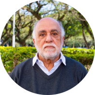 Foto de homem branco e idoso em ambiente aberto. Ele é calvo, tem olhos castanhos, cabelos curtos e brancos, barba branca, usa camisa azul e suéter azul escuro em decote V. Atrás dele há arbustos e árvores.