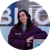 Foto de mulher branca sorrindo. Ela tem cabelos lisos, compridos e castanhos e usa uma blusa de frio roxa sobre outra blusa cinza. Ela tem um cotovelo apoiado sobre um móvel. Ao fundo, uma parede de vidro com adesivos de letras que formam parte da palavra biblioteca.