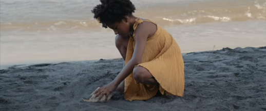 Foto mostra uma menina negra de cabelos crespos e escuros presos no alto da cabeça. Ela usa um vestido amarelo e está agachada sobre a areia da praia, enquanto segura uma grande concha. Ao fundo, é possível ver as ondas do mar quebrando.