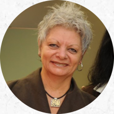 Foto de uma mulher branca de cabelos curtos e grisalhos. Ela usa brincos de argola prateados, um blazer marrom e um colar preto com pingente quadrado. Ela sorri e olha para a câmera. 