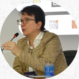 Foto de uma mulher de pele branca, cabelo curto escuro falando enquanto segura um microfone de mesa. Ela veste um sobretudo bege e usa óculos. Atrás dela há uma pequena parte visível de um telão com uma apresentação.