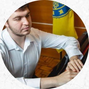 Foto de um homem em frente a um piano. O homem tem pele branca, cabelo curto e claro, barba e veste uma camisa branca. Ele está sentado em frente a um piano escuro com suas duas mãos em cima do instrumento. Seu olhar é direcionado ao piano. Parte de uma bandeira amarela com elementos azuis está posicionada ao fundo.