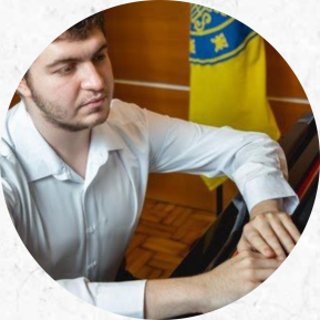 Foto de um homem em frente a um piano. O homem tem pele branca, cabelo curto e claro, barba e veste uma camisa branca. Ele está sentado em frente a um piano escuro com suas duas mãos em cima do instrumento. Seu olhar é direcionado ao piano. Parte de uma bandeira amarela com elementos azuis está posicionada ao fundo. 