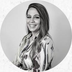 Clotilde Perez é uma mulher loira, com cabelos longos e, na foto (em preto e branco), está em pé e veste uma blusa estampada.