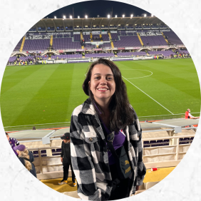 Foto de uma mulher na arquibancada de um estádio de futebol. Ela tem pele branca, cabelo e olhos escuros. Ela veste um casaco quadriculado em preto e branco e uma camisa roxa. Ela sorri e posa para a câmera. Atrás dela há um campo de futebol e parte da arquibancada com algumas pessoas.