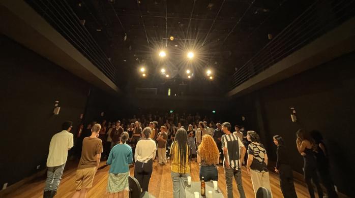 Foto da Mostra USP de Teatro Universitário em 2023. À frente, há 11 pessoas de pé, em cima de um palco de chão de madeira e de frente para uma plateia que também está de pé. As paredes e o teto são pretos. No teto também há uma iluminação forte com cinco lâmpadas amarelas.