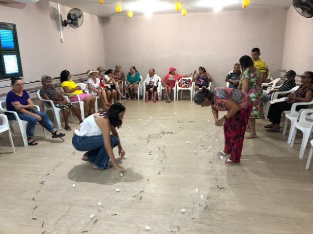Foto de um grupo de cerca de 20 pessoas, em sua maioria mulheres idosas, reunido em um semi-círculo, sentados em cadeiras de plástico dentro de uma sala. Ao centro, uma senhora e uma mulher um pouco mais jovem estão abaixadas mexendo em uma rede de pesca.