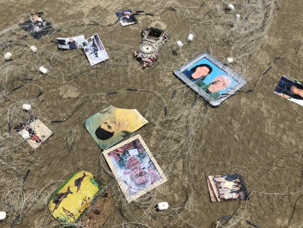Foto de uma rede de pesca na areia com alguns objetos dispostos sobre ela, como um relógio, algumas fotografias coloridas e em preto e branco e dois retratos pintados.