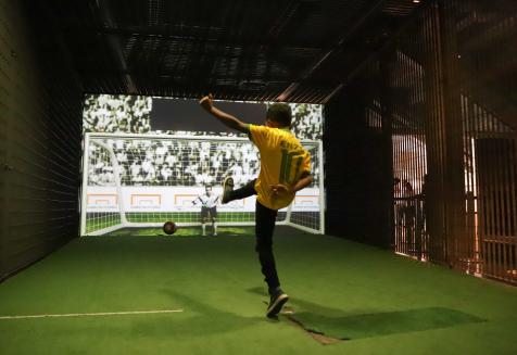 Foto de um menino negro com uma camisa do Brasil chutando uma bola de futebol em direção a uma parede com uma projeção da imagem de um gol, um goleiro e torcida. O chão é verde. Na camisa, está escrito o nome Enzo em cima do número 10. A criança está com um braço atrás do corpo e o outro estendido para cima. Uma perna está no chão, nas pontas do pé, e a outra está estendida em 90º graus, na mesma direção da bola.