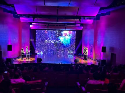 Foto do palco durante a premiação. No telão ao fundo, está sendo projetada uma imagem de galáxia com o texto “indicados” e o logo da USP em branco. Em cada extremidade do palco, há duas pessoas em pé. A iluminação está baixa e luzes roxas estão direcionadas para as paredes. Na porção inferior da imagem, está o público sentado assistindo.