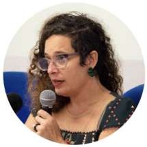 Foto de mulher branca em semi-perfil falando ao microfone. Ela tem cabelos castanhos cacheados e compridos com a lateral mais curta, usa óculos, brinco verde e uma blusa verde escura com bolinhas vermelhas.