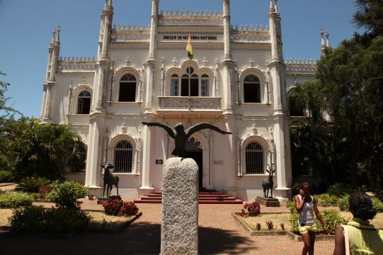 Foto do Museu de História Natural, ponto turístico de Maputo. No centro da imagem há um edifício com estrutura gótica, de cor bege com torres e janelas ornamentadas. Algumas árvores estão à frente e ao lado do prédio. No primeiro plano, ao centro, há a estátua de um pássaro com as asas abertas sobre uma pedra. No canto direito, duas mulheres caminham.