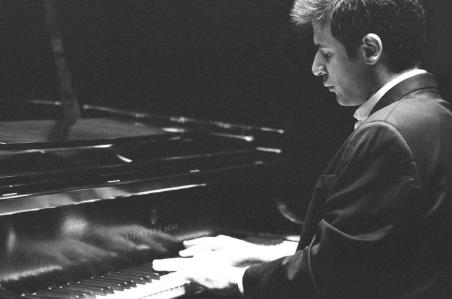 Foto em preto e branco. Cristian Budu é um homem branco, de cabelos curtos, lisos e escuros. Na foto, ele está de lado, sentado em frente a um piano, com as mãos sobre as teclas. Veste um terno preto.