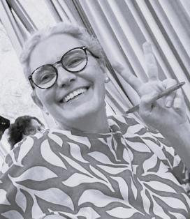 Foto em preto e branco de um mulher branca, cabelos grisalhos, que estão presos. Ela usa oculos de hastes escuras e arredondadas. Veste uma camisa estampada. Ela olha para a camera e sorri, fazendo sinal de V com uma das mãos.