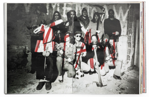 Foto de um livro aberto, visto de cima. Ele exibe, dividida em duas páginas, a foto em preto e branco de um grupo de aproximadamente 10 pessoas. A maioria são homens brancos que estão agachados ou em pé na frente de uma parede com pichações. Por cima da foto, está anotado em vermelho, em letras grandes, “M I A W”.