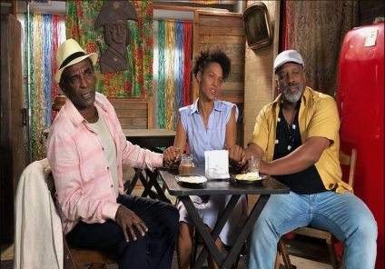 Foto de uma cena do filme O Pai da Rita. Três pessoas estão sentadas em uma mesa de bar. Nas pontas estão dois homens negros: na esquerda, um deles veste uma camisa rosa claro xadrez por cima de uma camiseta branca e uma calça preta, além de um chapéu. Na ponta direita, um homem de boina e barba grisalha veste uma camisa mostarda por cima de uma polo preta e uma calça jeans. No meio, está uma mulher negra segurando a mão dos dois homens. Ela usa um vestido azul claro e tem cabelos curtos e crespos.