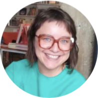 Foto de mulher branca sorrindo. Ela tem cabelos castanhos claros e lisos até os ombros e olhos claros. Ela usa óculos de armação quadrada e avermelhada e veste blusa verde. Atrás dela, uma sala com mesa e prateleiras.