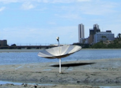 Foto de uma antena parabólica fincada na lama e na areia em meio à água. No fundo, o céu azul e prédios e árvores no horizonte e, no fundo, o céu azul.