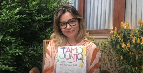 Patrícia Guimarães Gil é uma mulher jovem, de cabelos loiros na altura dos ombros e óculos de armação grande e escura. Ela usa uma blusa estampada em tons de marrom e segura seu livro Tamo Junto: oargumento estudantil e sua gramática em uma arena de conflitos enquanto sorri sutilmente para a câmera. 