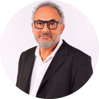 Foto de homem branco de olhos castanhos, cabelos e barba curtos e grisalhos. Ele usa camisa branca sob blazer preto e óculos de armação retangular e preta. O fundo é claro.