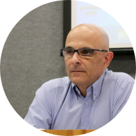 Foto de homem branco e careca olhando para a esquerda da imagem. Ele tem olhos castanhos, veste camisa social azul clara e usa óculos de armação quadrada e preta. Ao fundo, uma parede cinza e parte de uma tela de projeção.