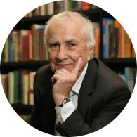 Foto de um homem idoso branco, com olhos escuros, cabelos curtos, lisos e brancos, usando uma camisa branca sob um terno preto. Ele posa para a foto apoiando o queixo sobre a mão. Ao fundo, há uma estante repleta de livros. 