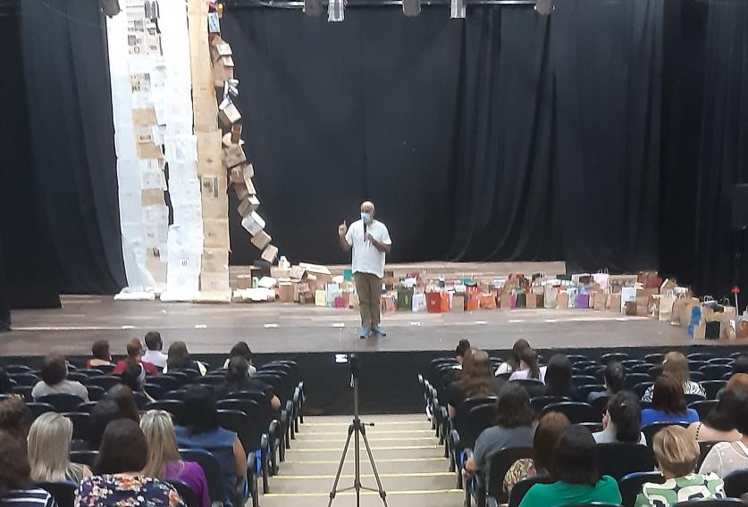 Foto tirada no fundo de um auditório. Algumas pessoas estão sentadas olhando para o palco, onde está Wilton, que é branco, careca, usa máscara e veste uma blusa branca. Atrás dele, está a obra de arte, que é composta por uma espécie de cortina, formada por papéis brancos e beges. Do topo da cortina, saem algumas caixas de papelão ligadas em fila até chegar no chão, onde estão agrupadas diversas sacolas de papel. 