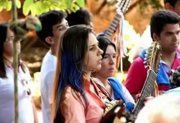 Foto de Priscila Maria Ribeiro durante a folia de reis. Ela é uma moça de pele branca e madeixas longas com mechas azuis e toca uma viola caipira rodeada de pessoas vestidas à caráter para a festa do reisado.