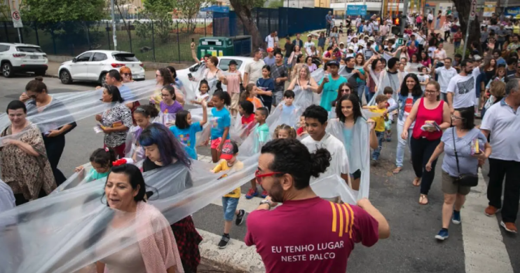 Foto de diversas pessoas em um cortejo na rua. São mulheres, homens, crianças e adolescentes, de diferentes tons de pele. Muitas pessoas estão conectadas por uma faixa extensa de plástico transparente encaixado em seus pescoços, formando filas dentro do cortejo. Na parte inferior da foto, destaca-se um jovem branco de barba e cabelos escuros e cacheados presos. Ele está de costas, usa um óculos de armação vermelha e uma camiseta com os dizeres “eu tenho lugar neste palco”. 